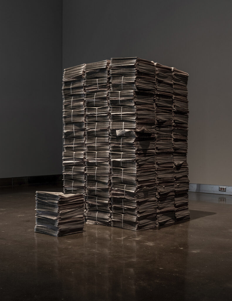 Very large stack of bundled papers in a dimly lit gallery space.