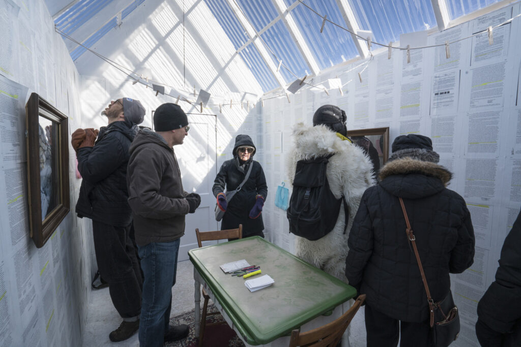 Five people in winter clothing viewing text and art inside a white shelter with a clear roof.