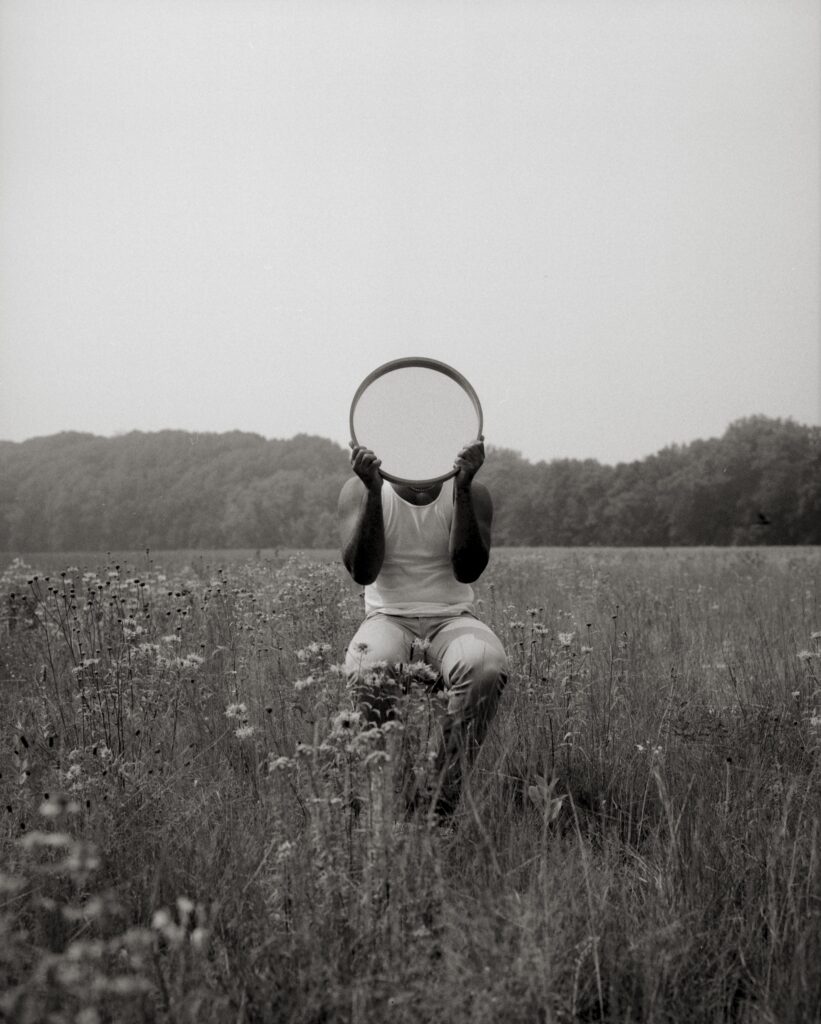 Black and white photograph of a person with dark skin sitting in a prairie holding a round mirror over their head.