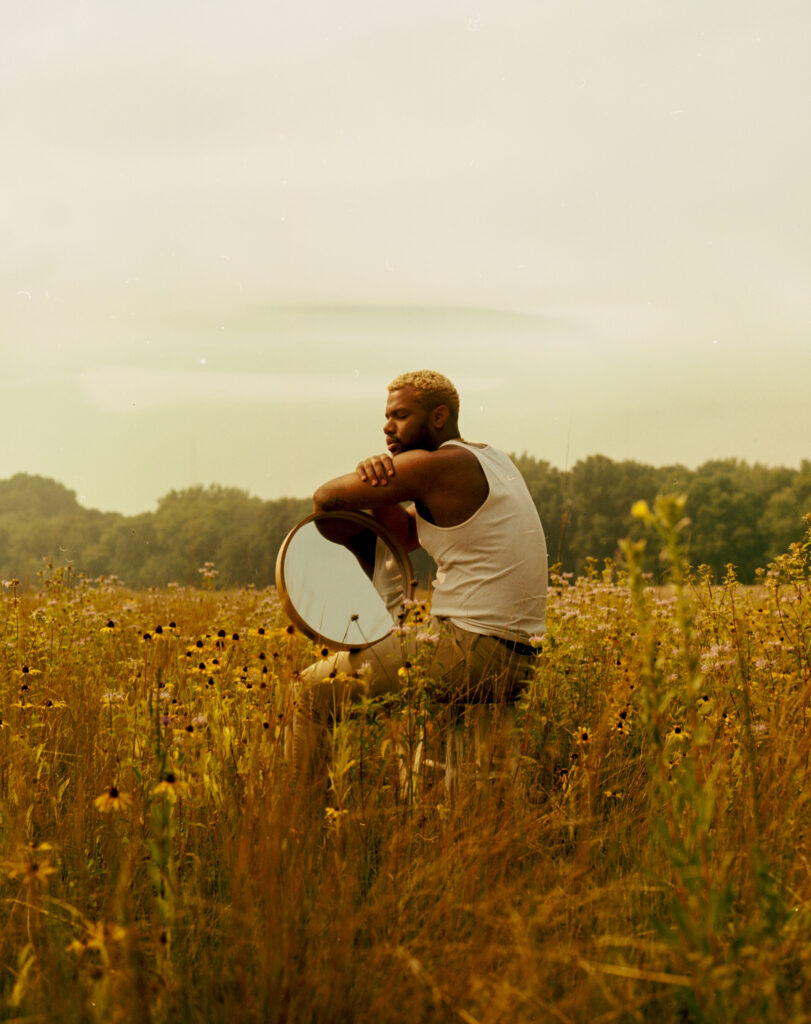 Photo of a man with dark skin sitting in a prairie, resting a mirror under his arms.