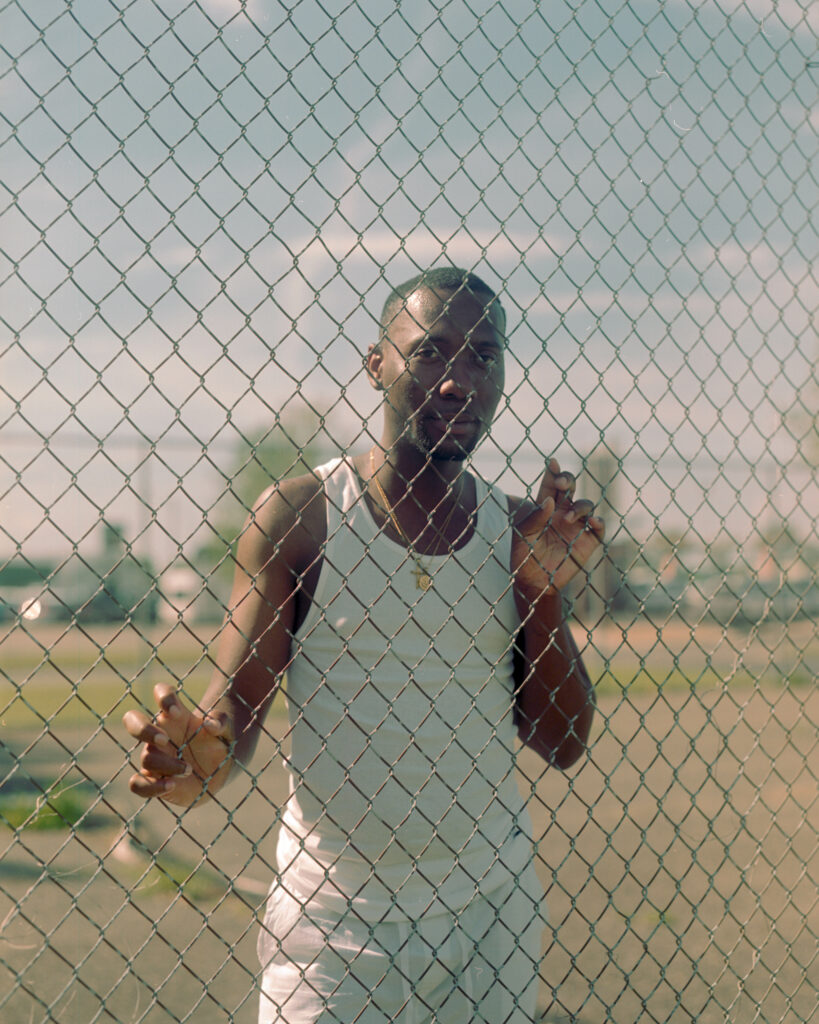 Photo of a man with dark skin wearing white and standing behind a chainlink fence.