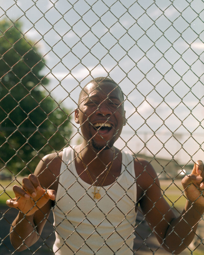 Photo of a man with dark skin standing behind a chainlink fence yelling.