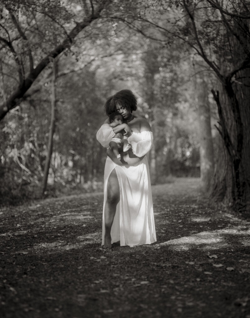 Black and white photo of a woman with dark skin holding a baby and standing in a forest path.