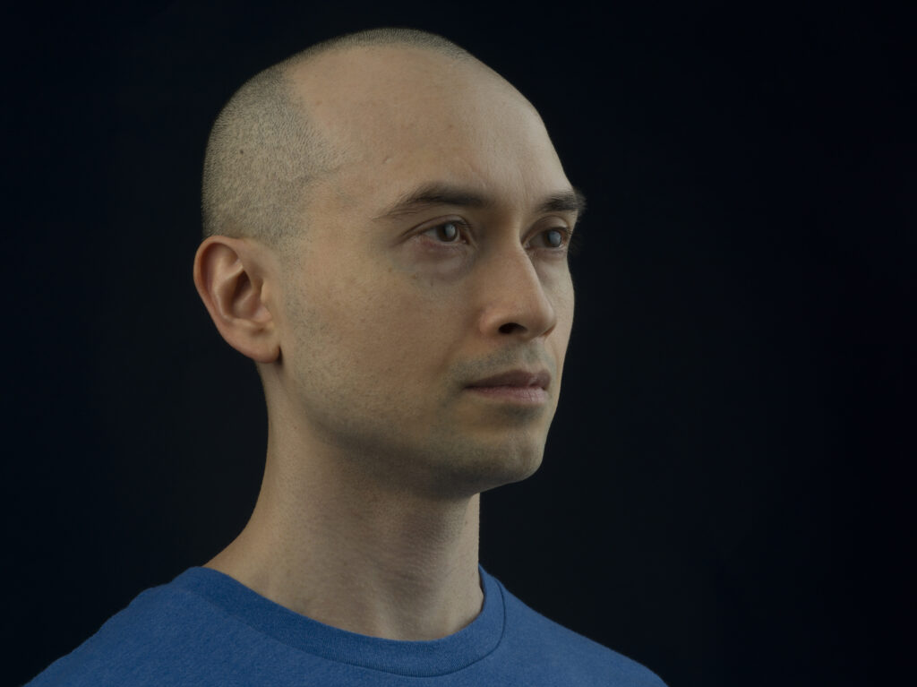 Angled portrait of a person with a shaved head staring away against a black background.