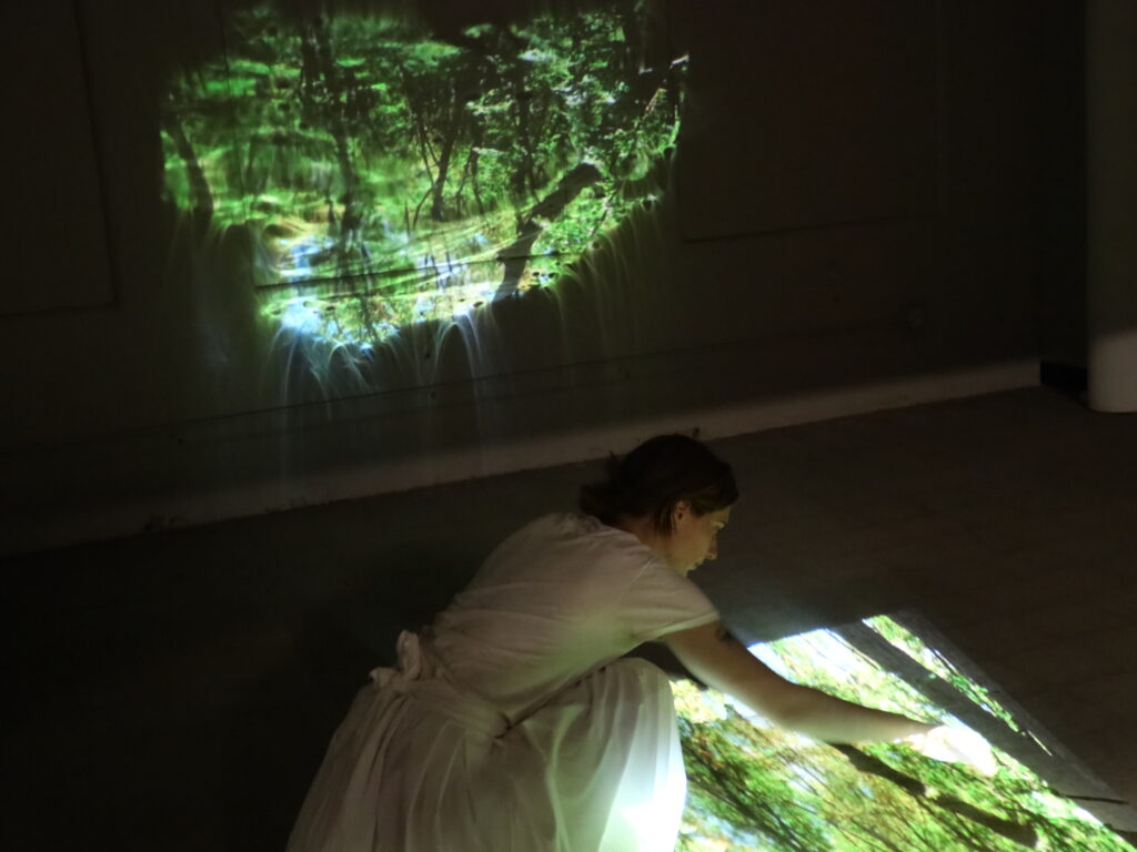 Person wearing white dress kneels and touches projected image of green forest on floor, with mirrored projection on wall behind them.