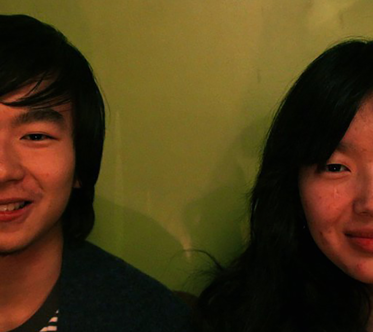 Two people with dark hair smile at the camera, green wall behind them and the outside halves of their faces cropped from the frame.