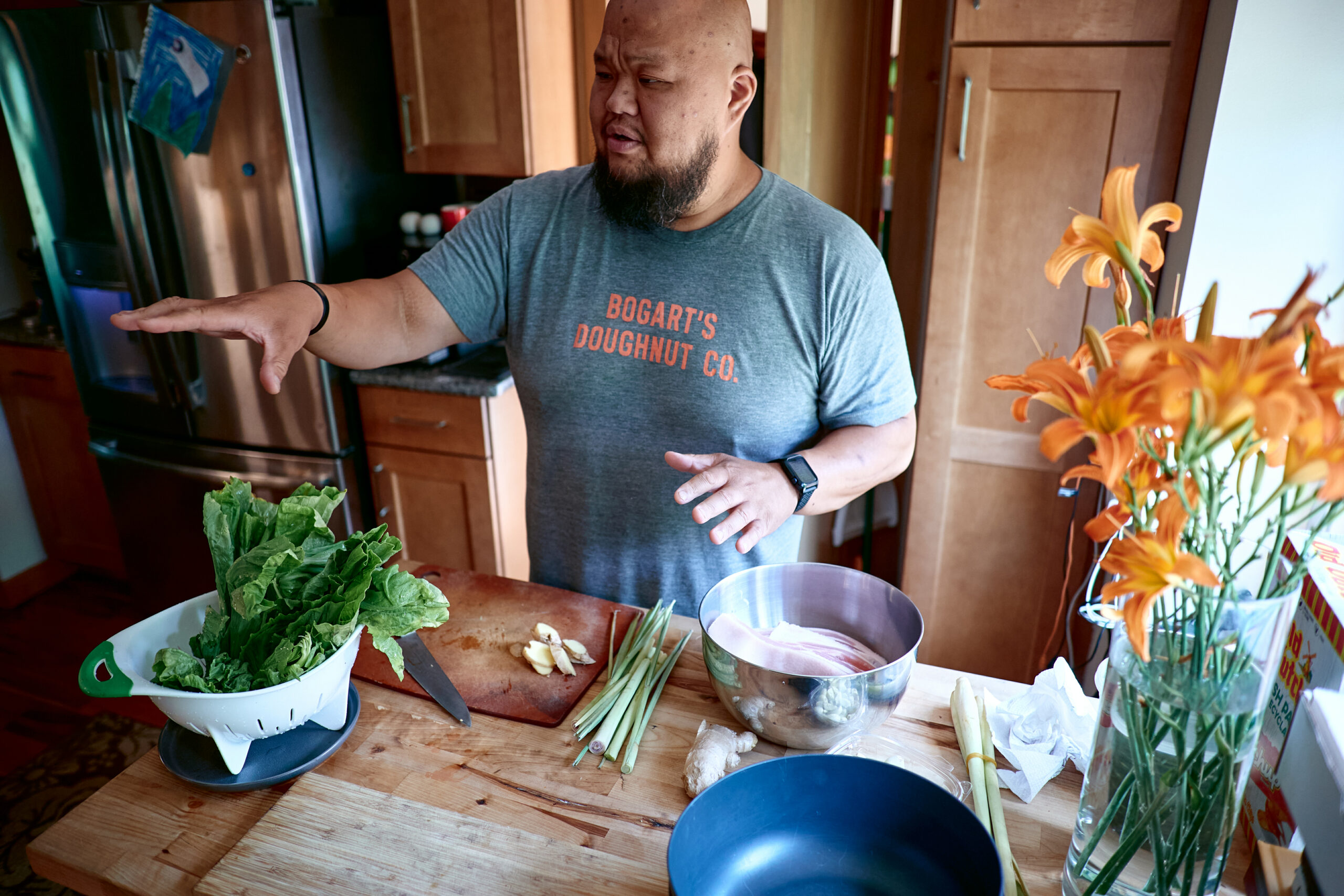 Yia Vang wears gray Bogart's Doughnut Co. t-shirt and gestures in front of kitchen island with many ingredients.