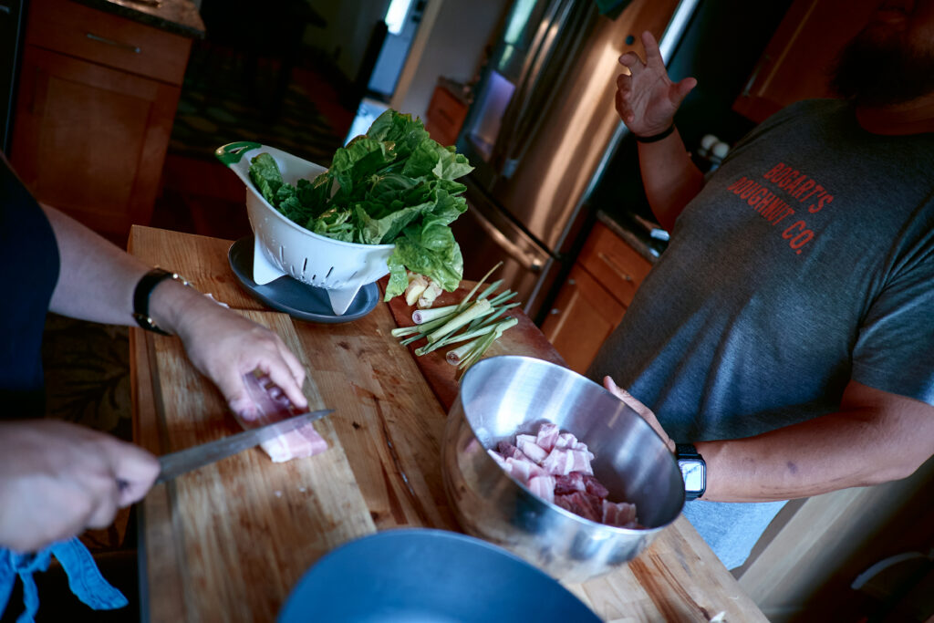 Hands cutting pork on kitchen island, with metal bowl and colander of greens, and person gesturing.