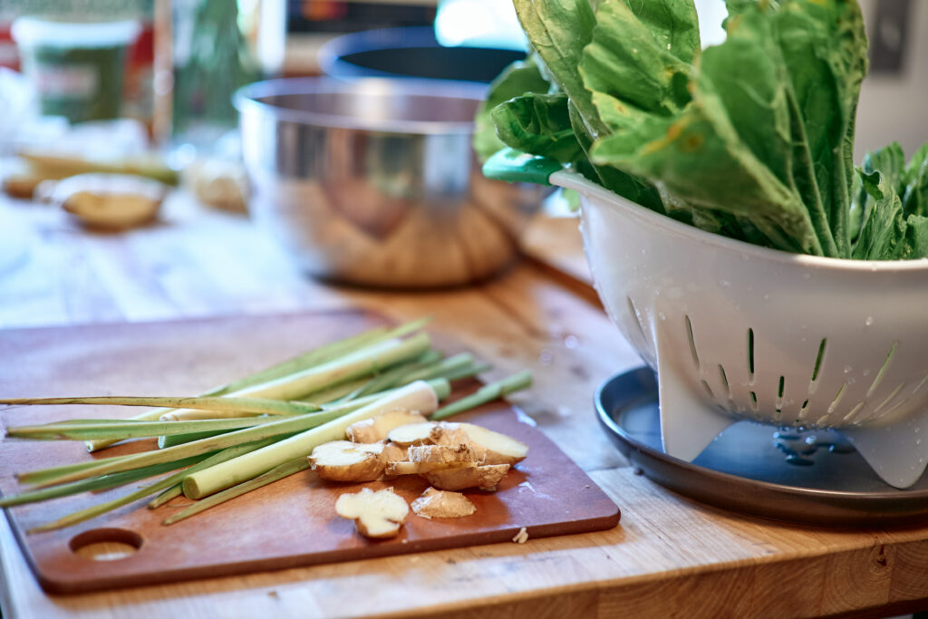 Lemongrass and ginger on cutting board, with colander full of greens beside and metal bowl behind.