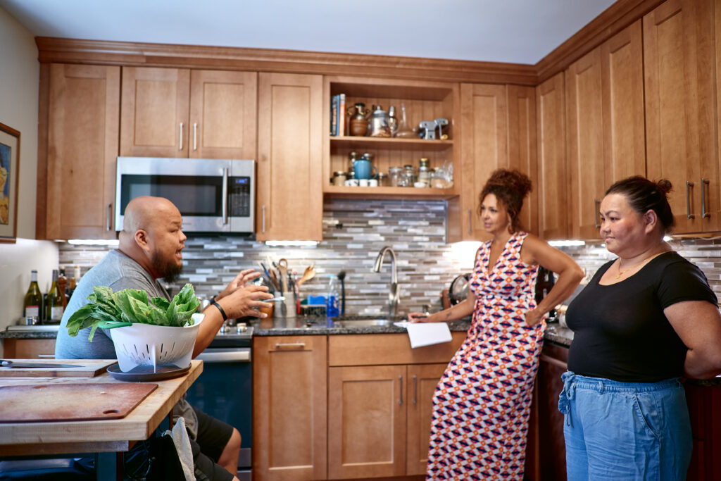 Three people stand talking in kitchen with wooden cabinets.