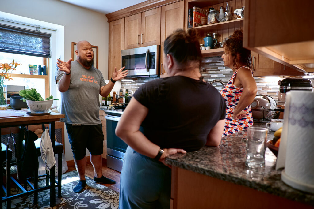 Three people stand talking in kitchen with wooden cabinets.