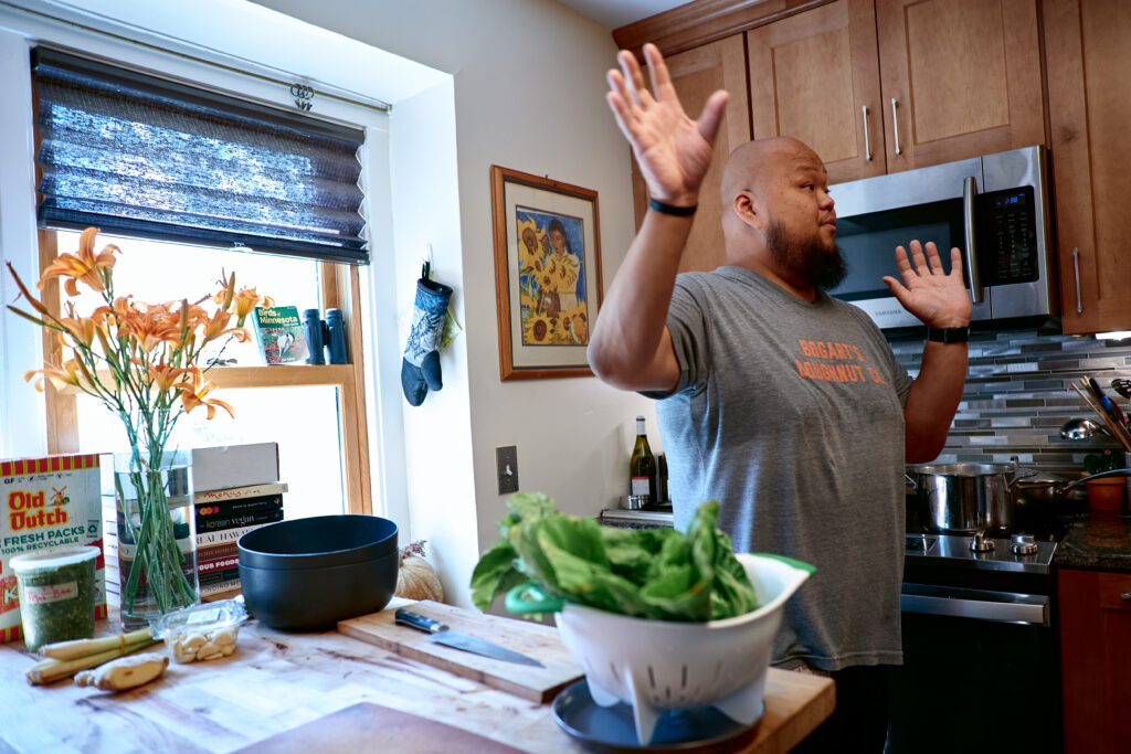 Yia Vang gestures with hands raised in front of stove, with colander on kitchen island in foreground.