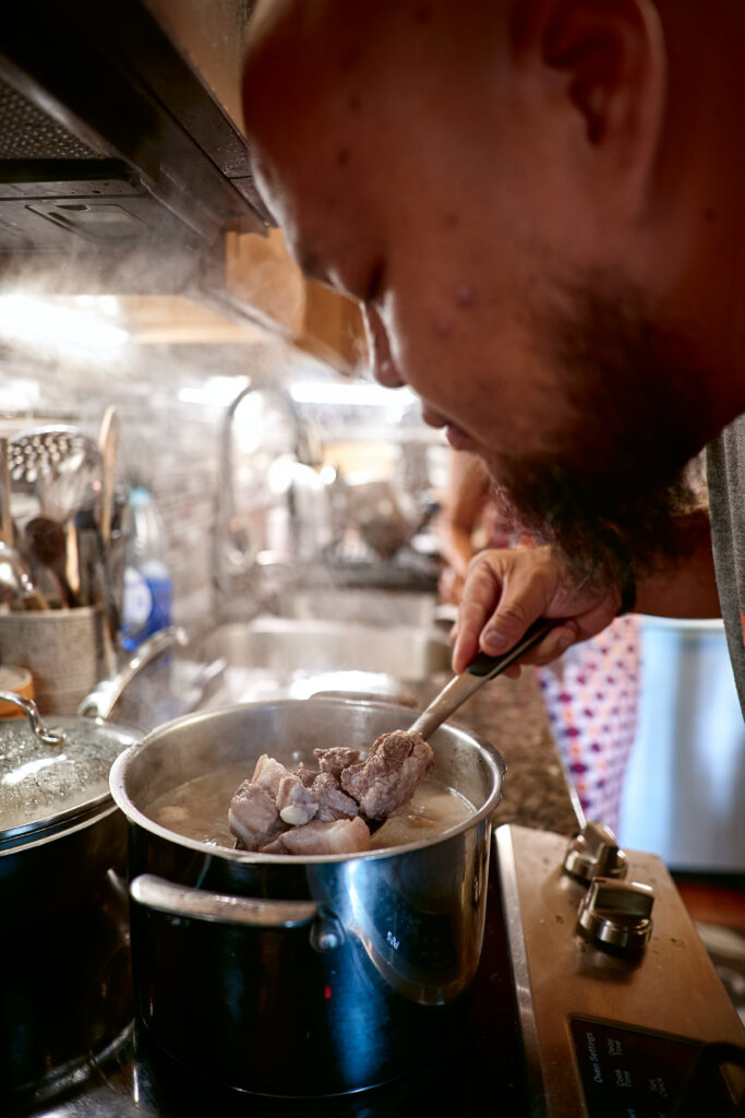 Yia Vang lifts spoonful of pork out of pot on stove.