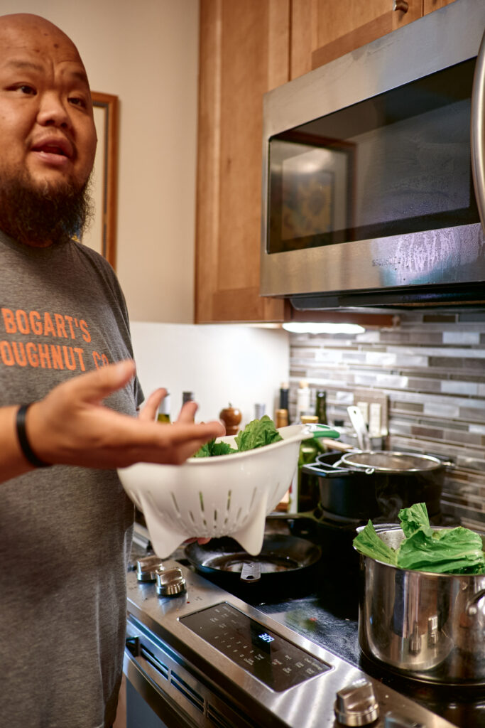 Yia Vang gestures in front of stovetop, holding colander of greens.