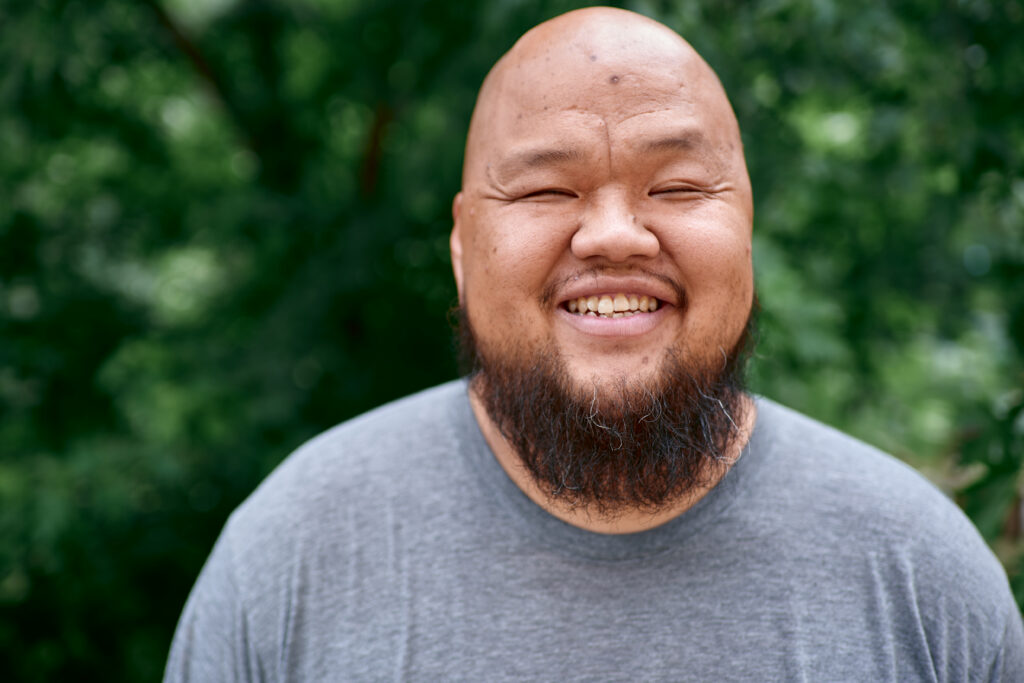 Hmong person with beard and gray t-shirt laughs with his eyes closed, green trees in background.