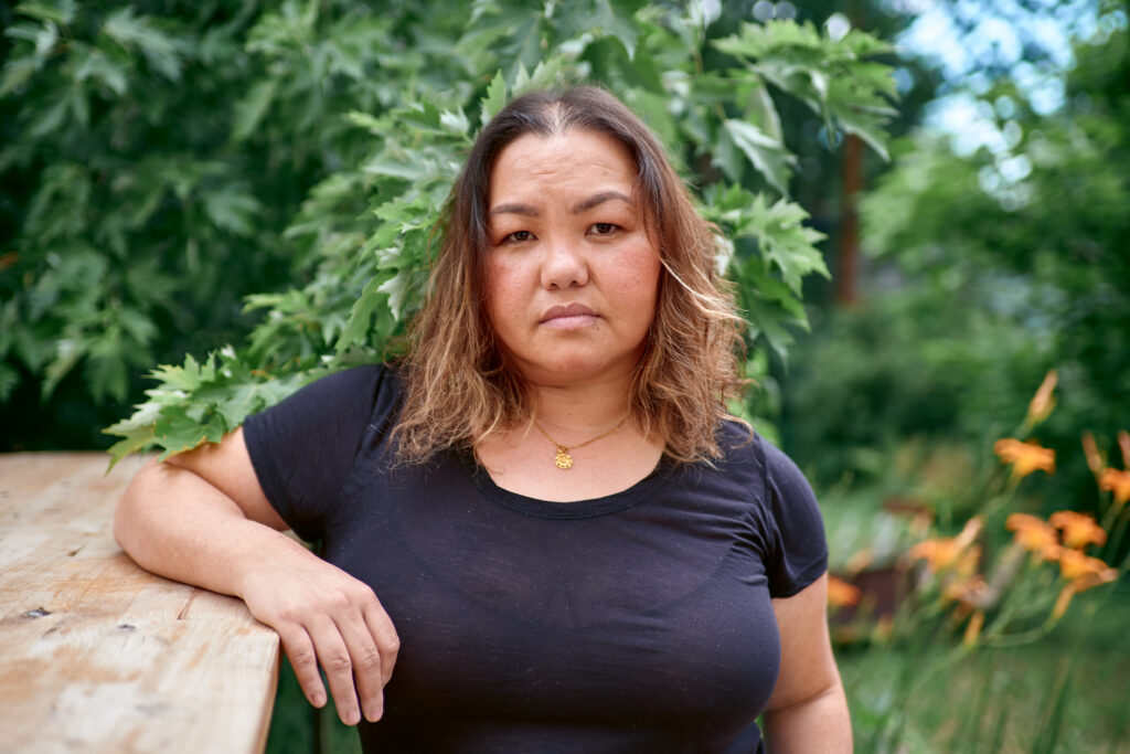 Hmong person with long hair wears black t-shirt and gold necklace, looks directly at the camera with her shoulder leaning on a railing, green leaves behind her.