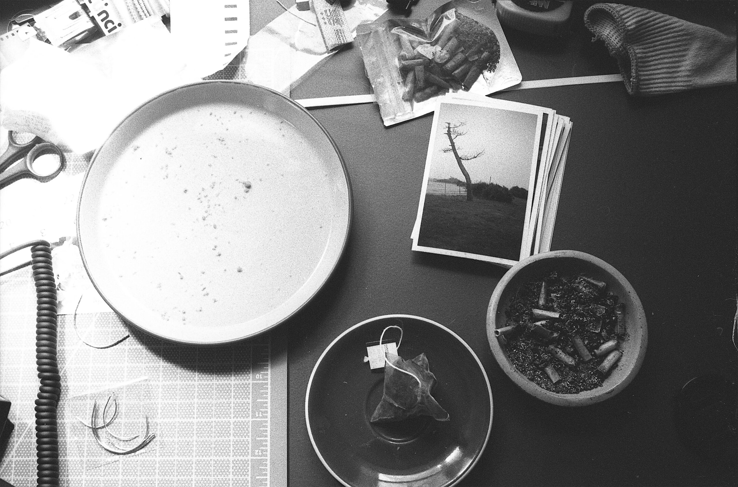 Black and white overhead photo of desktop with empty plate, used teabag, ashtray, photographs, and phone cord.