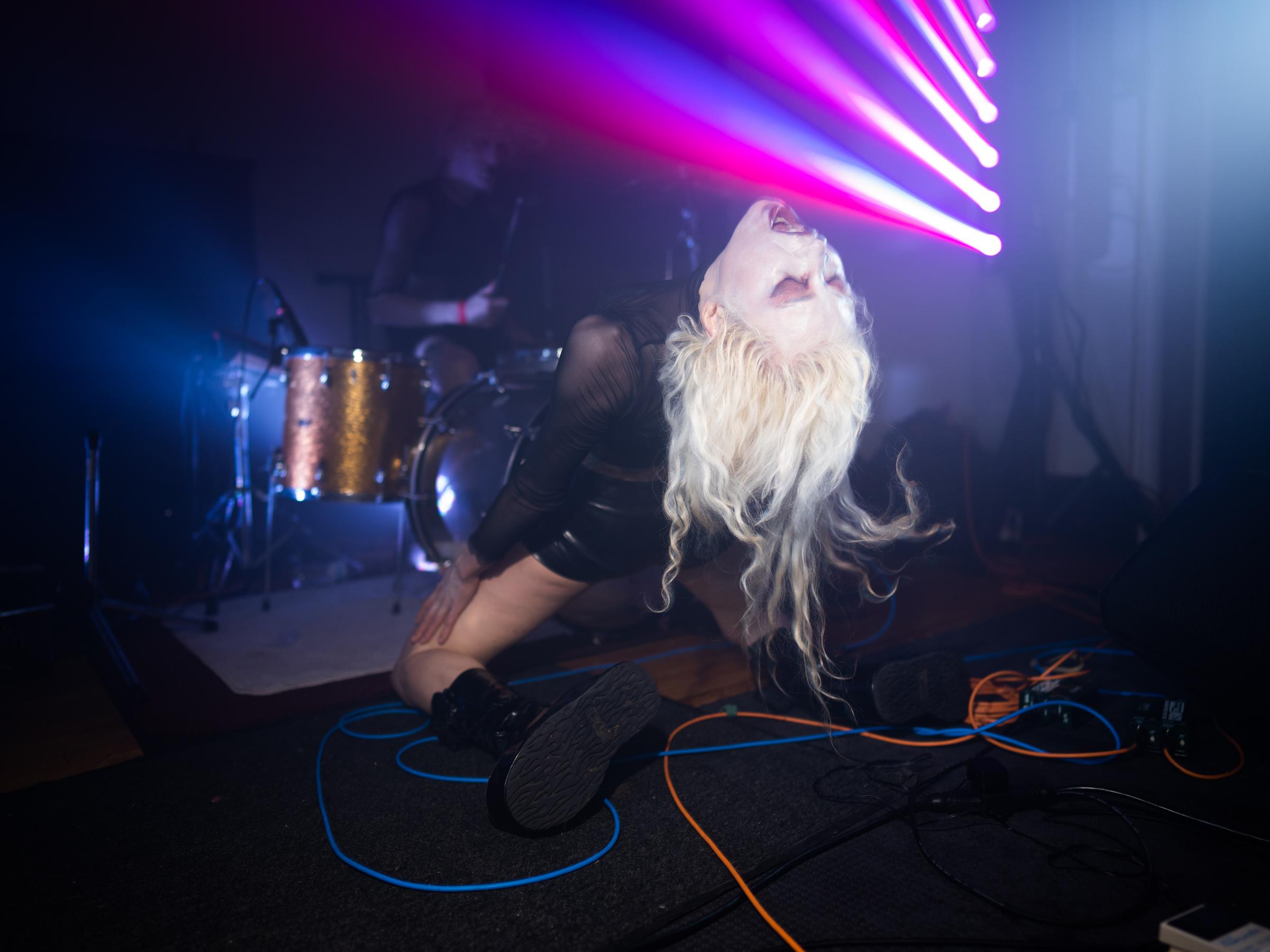 Performer with bleached blonde hair and white makeup kneels with head back in front of drum set, with purple and blue lighting above.