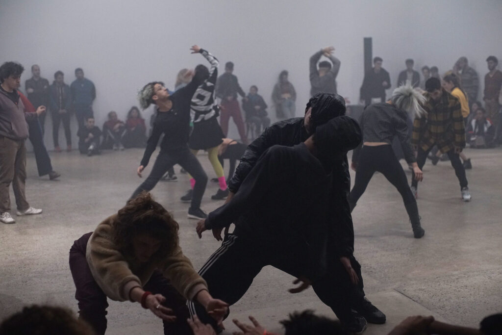 Many dancers in dark clothing, with gray theatrical fog and audience standing around edges of room.