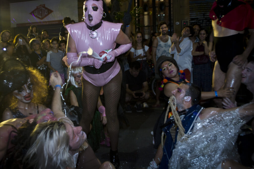 Performer wearing pink latex costume with hood and fishnets squirts white substance onto crowd.