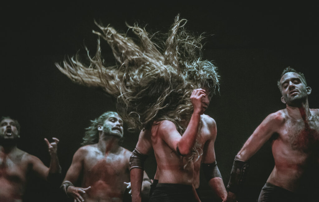 Group of performers with bare chests, and one performer in center with long brown hair flying in the air.