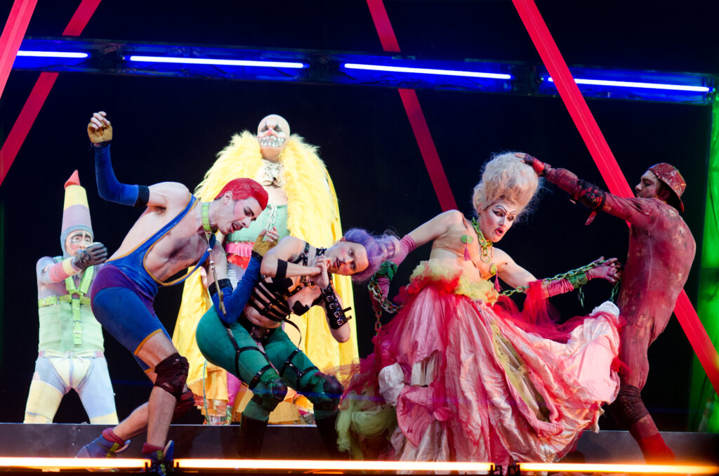 Performers in makeup and colorful costumes strike various poses surrounded by set pieces of colored light.