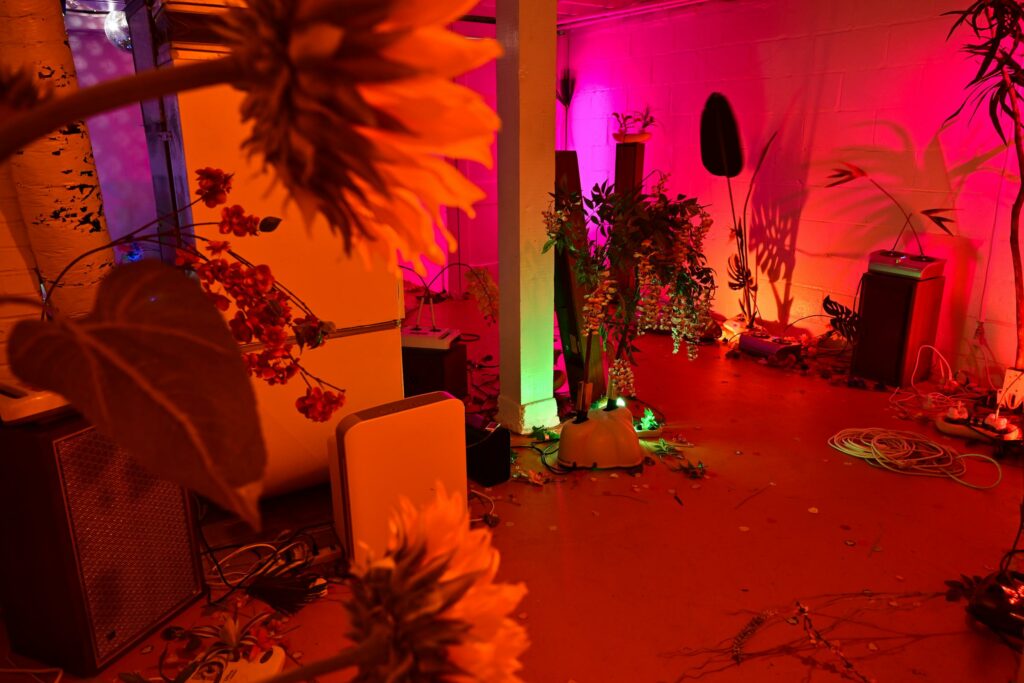 Gallery installation with fake flowers, household appliances, and orange and pink lighting.