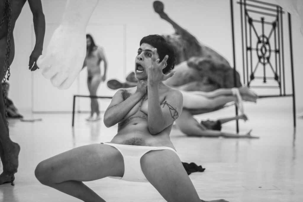 Performer wearing white underwear lunges with one hand open and the other at their throat, mouth wide.