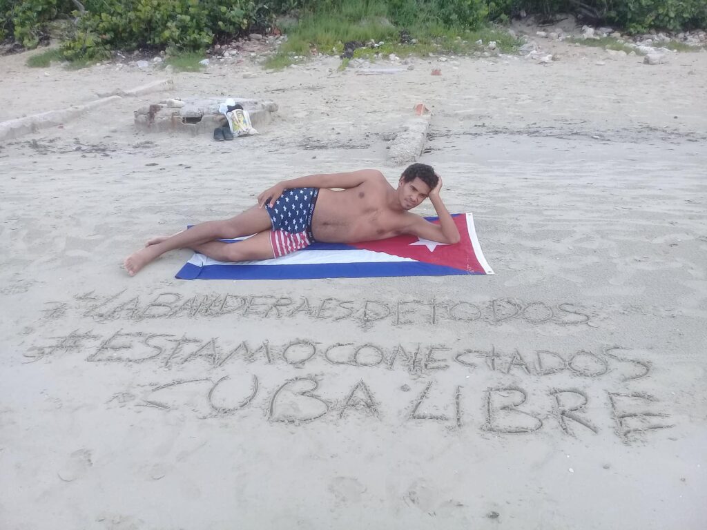 Person wearing USA flag print swimsuit lays on top of Cuban flag on beach, with writing in sand in front.