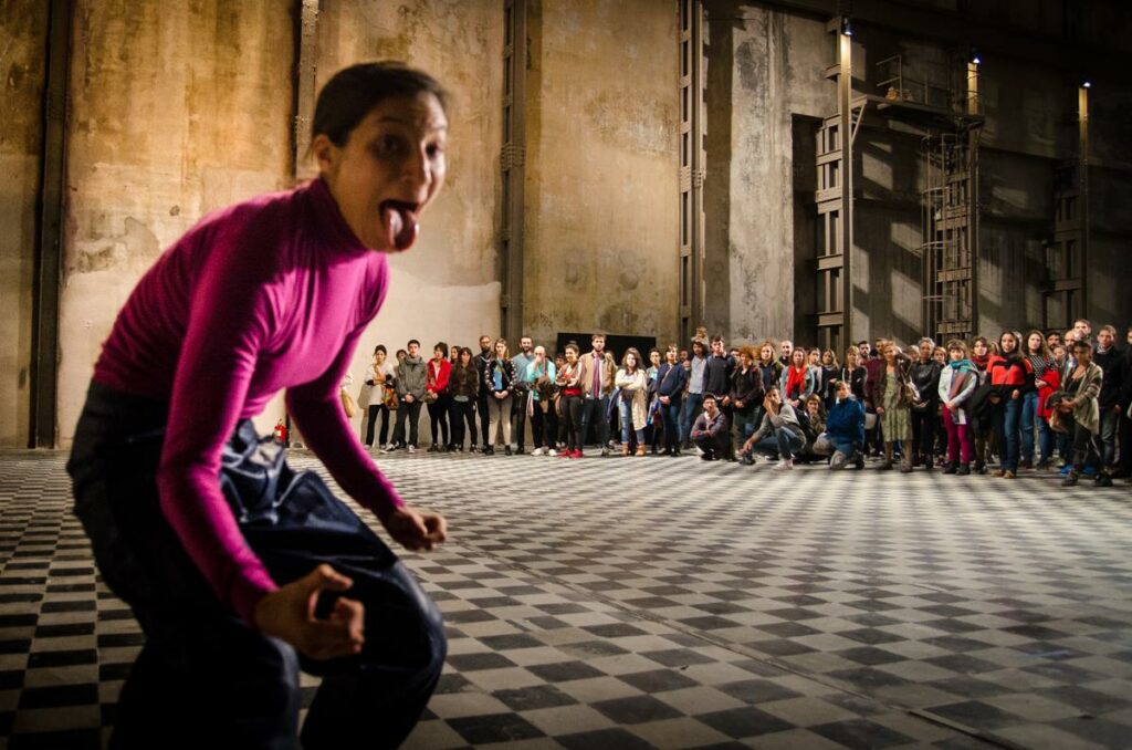Performer crouches with fists and tongue out, with large tile floor and audience gathered in background.