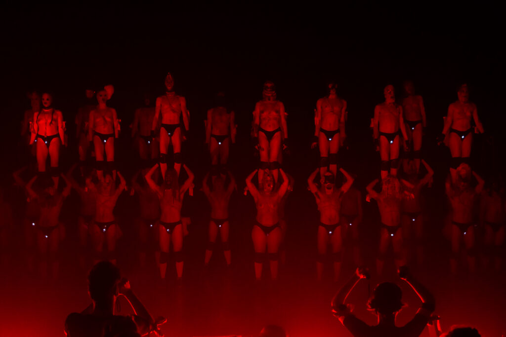 Row of performers standing on the shoulders of another row of performers, all bathed in red light.