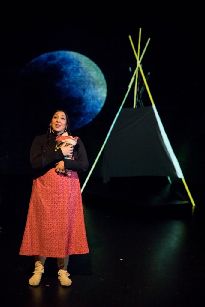 Actor wearing braids and long red dress in front of tent-like set design and projection of moon.