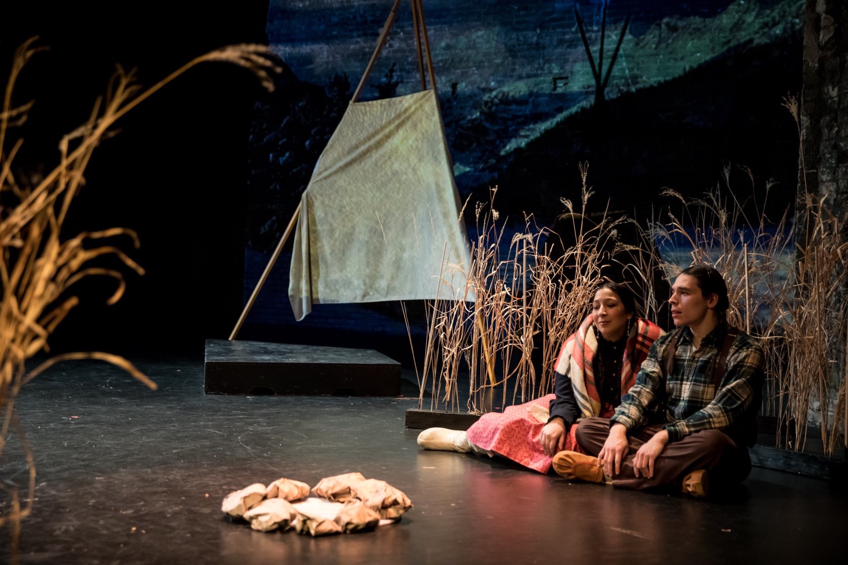 Two actors sit next to each other onstage, with set design including stalks of wheat and blue projection.