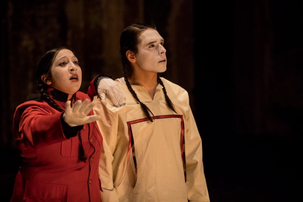 Two actors wearing braids and face makeup, one with outstretched hand.