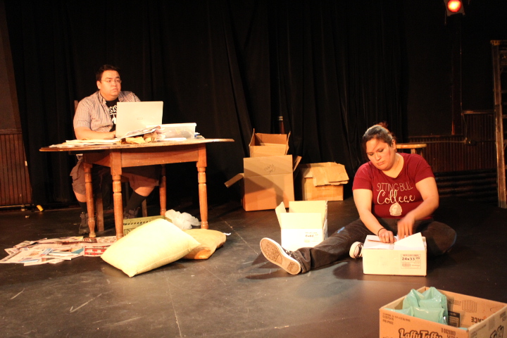 Two actors onstage, one sitting behind a desk and the other sitting on the floor.