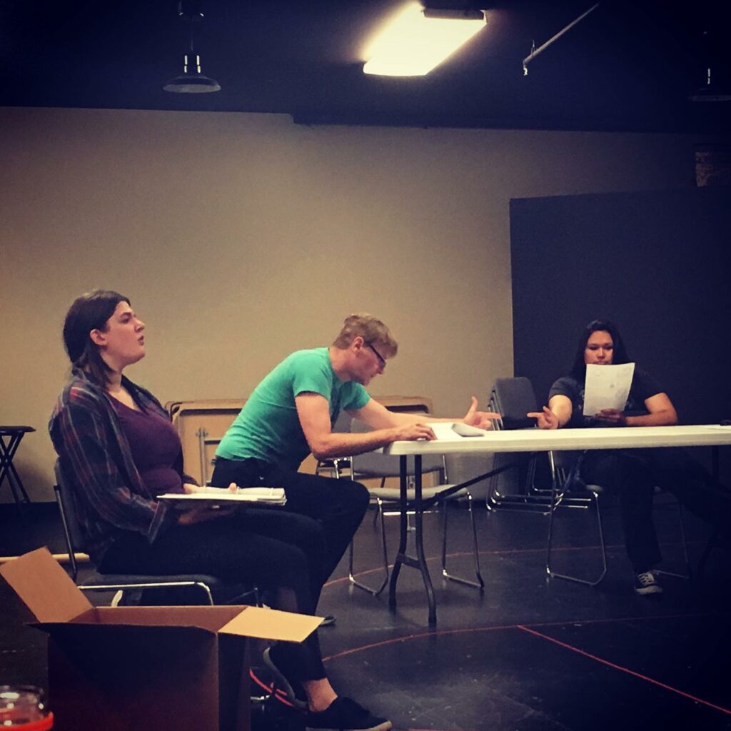 Three actors in rehearsal room, holding scripts around a folding table.