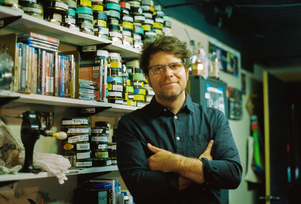 Person with light skin, brown hair, and glasses crosses arms in front of shelves of film reels.