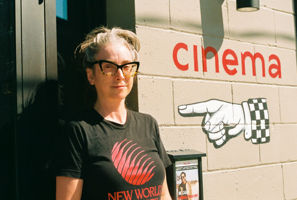 Person with gray hair in ponytail and cat eye glasses stands in front of wall painted with "cinema" and a hand pointing left.
