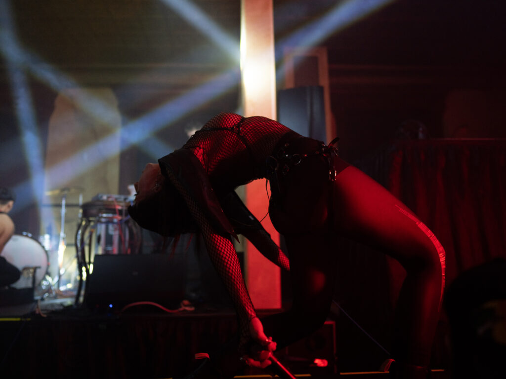 Performer wearing fishnets does a deep backbend in low red light, with beams of white light crossing behind them.