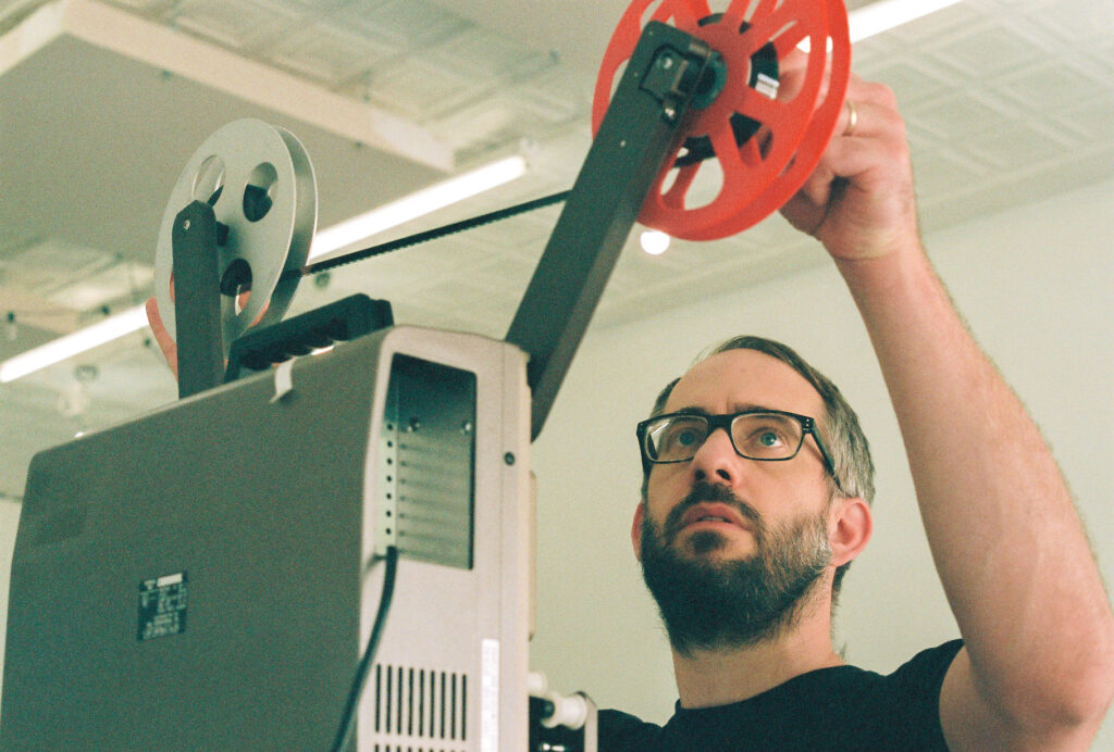 Person with beard and glasses operates 16mm film projector.