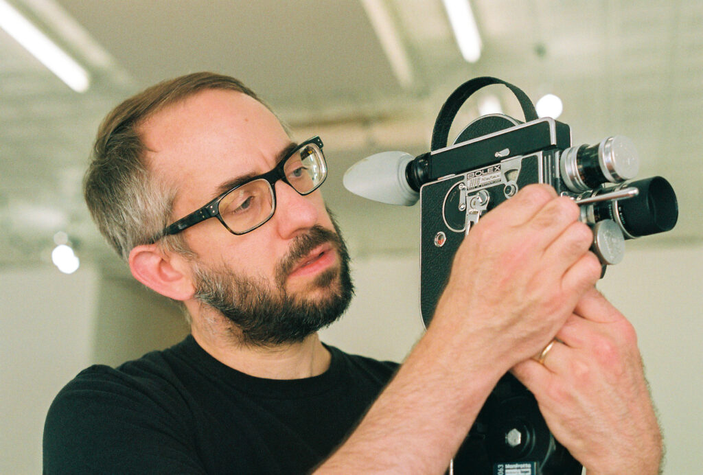 Person with beard and glasses holds Bolex camera.