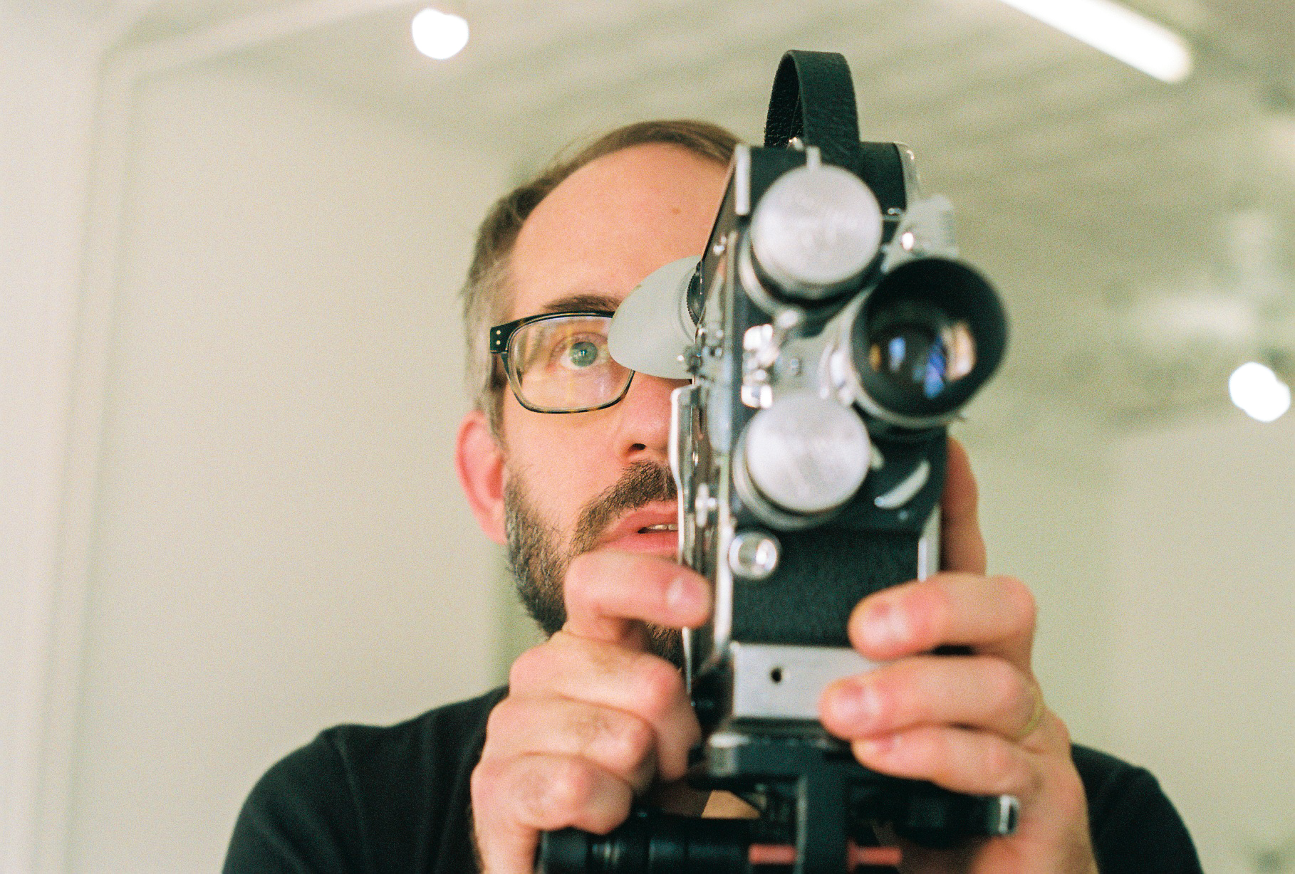 Person wearing glasses looks into the viewfinder of a Bolex camera pointed at the viewer.