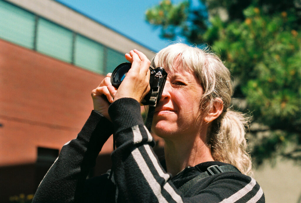 Person with white hair in ponytail holds film camera to their face and adjusts the lens.