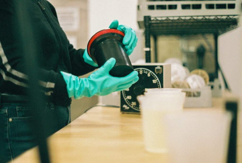 Hands wearing teal rubber gloves pick up black and red canister, with timer in background.