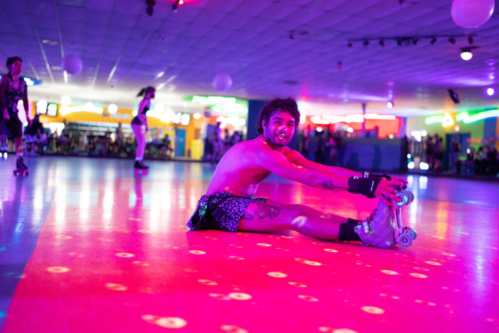 Person sits on skating rink and reaches down to grab their roller skates, with purple and pink light.