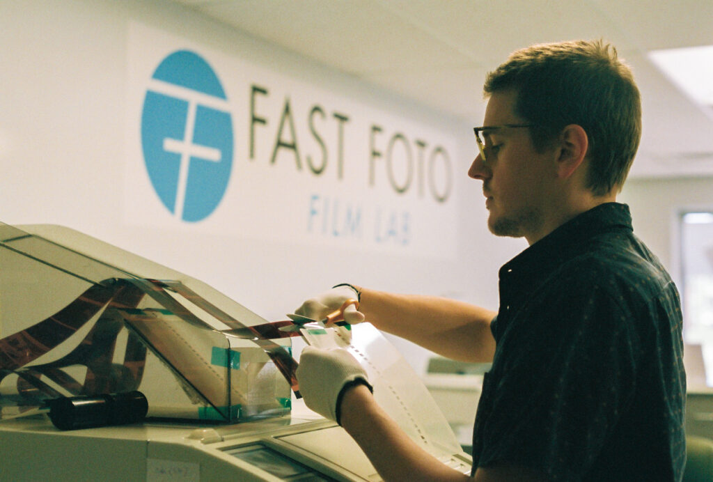 Person feeds film strip into machine, with Fast Foto and blue logo on wall behind.