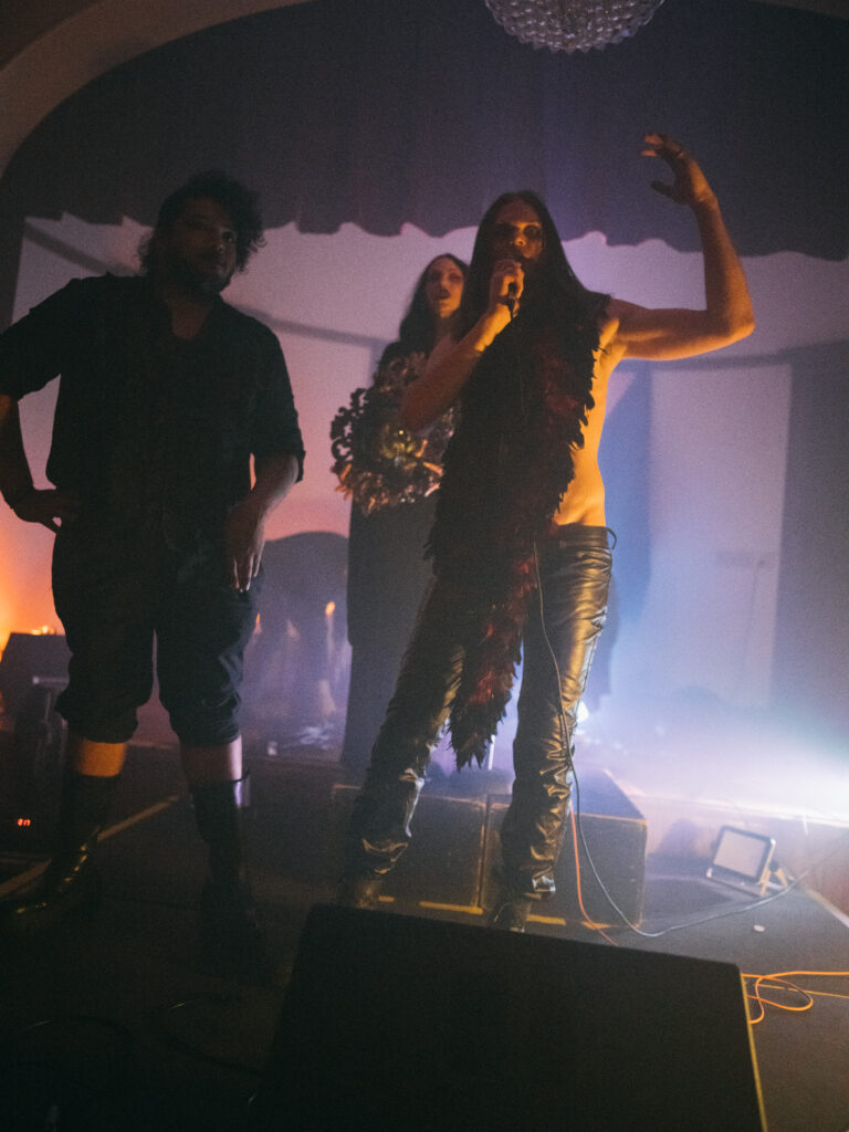 Person wearing leather pants speaks into microphone on stage with two people behind them.