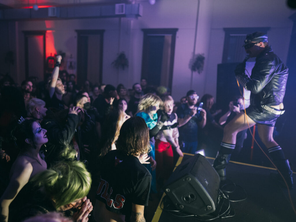 Performer onstage dressed in black leather, with large crowd in front of them.