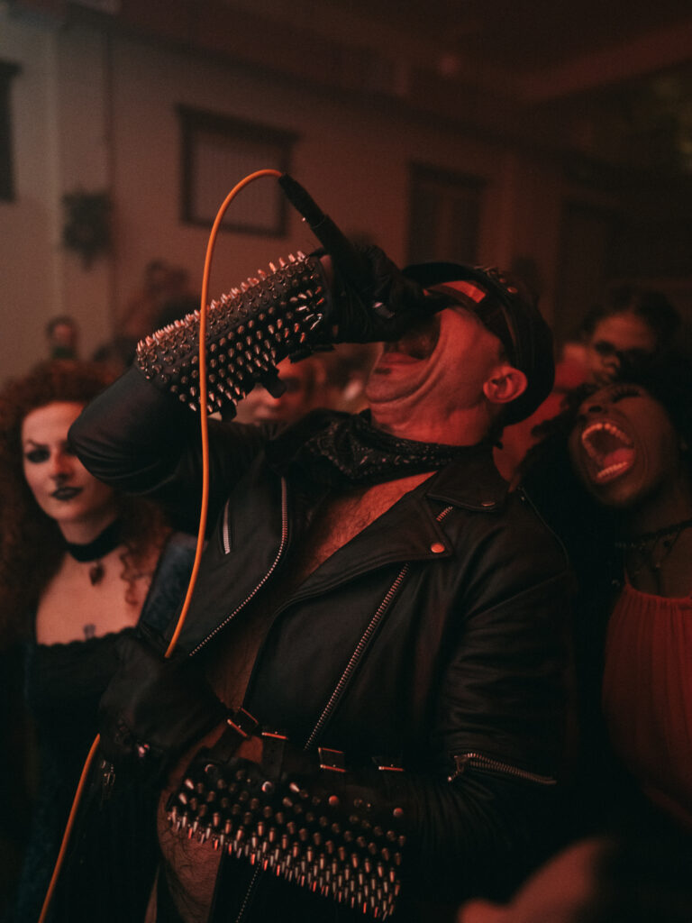 Performer wearing leather jacket sings into microphone, mouth open and head thrown back.