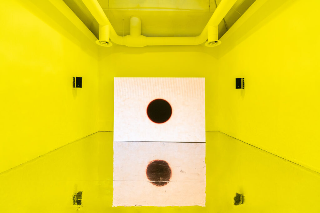 Bright yellow room with reflective floor, two speakers, and a screen at the back